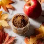 A jar of homemade Apple Pie Spice Mix surrounded by apples and cinnamon sticks.