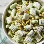 Bowl of homemade pistachio puppy chow with green and white cereal, nuts, and chocolate.